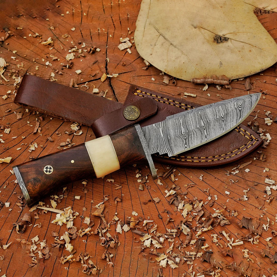 A hand forged Damascus steel hunting knife with a wooden handle and a leather sheath, resting on a wooden surface with wood shavings around.