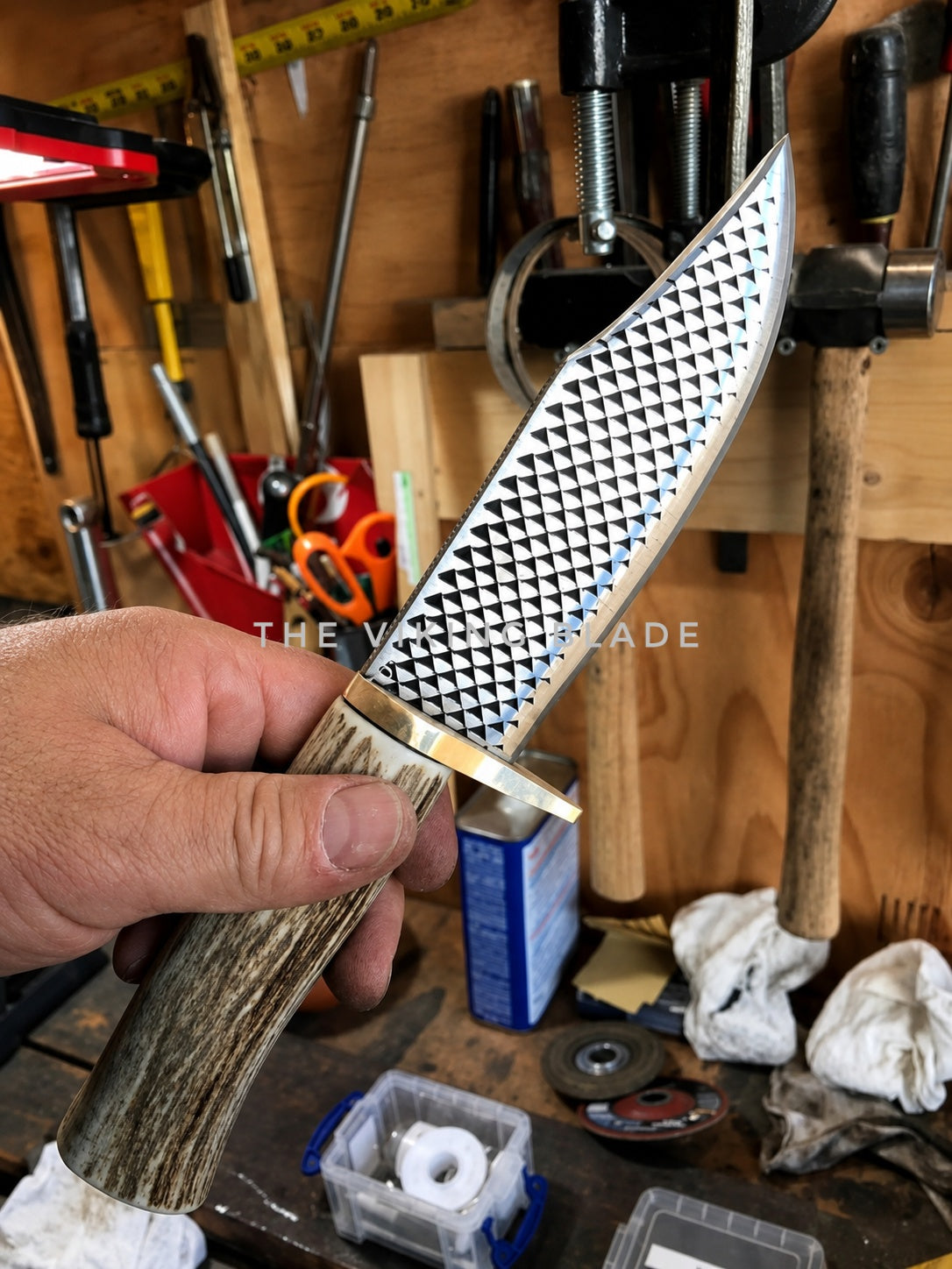 A hand holds a custom knife with a hammered silver blade and a textured brown antler handle in a workshop setting with tools in the background.