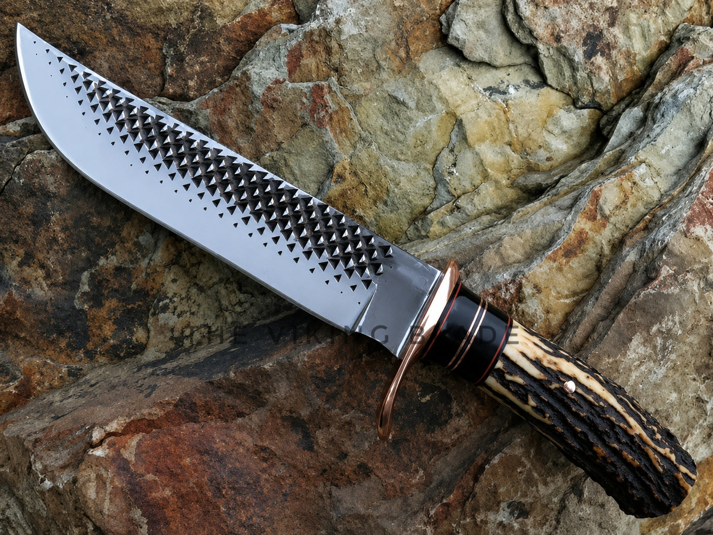 A fixed-blade knife with a textured, spike-patterned silver blade, a copper-colored guard, and a carved handle made of natural antler-like material, resting on a textured brown and gray rock surface.