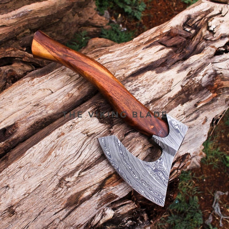 A hand-crafted Viking-style axe with a Damascus steel head and a curved wooden handle, resting on a textured log outdoors.