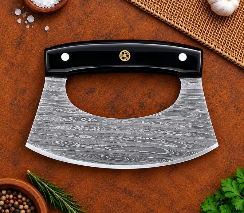 A kitchen cleaver-style knife with a patterned Damascus steel blade and a glossy black handle, displayed on a textured brown surface with rustic food props.