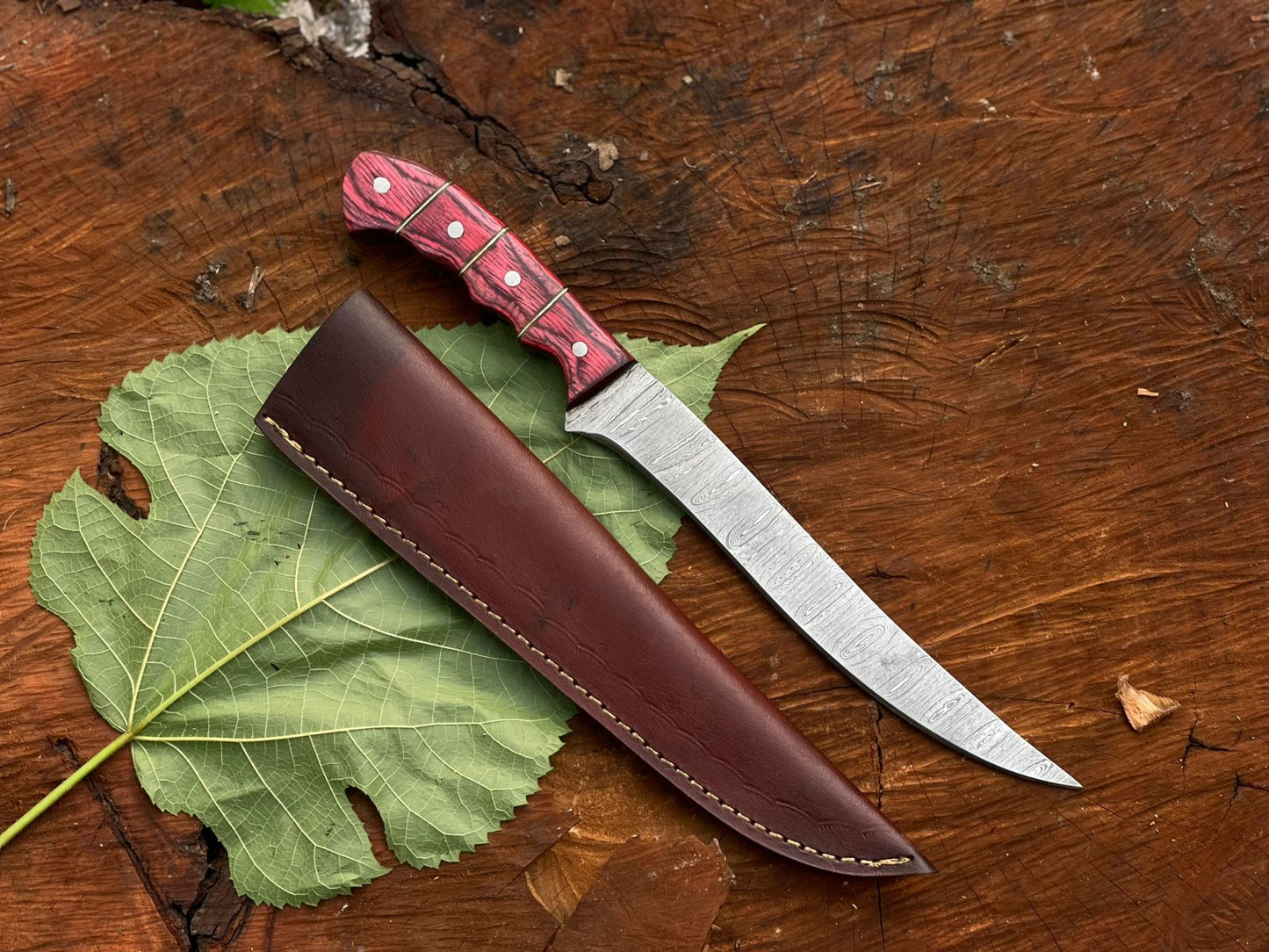 Knife with red handle and brown sheath on a wooden surface with a leaf