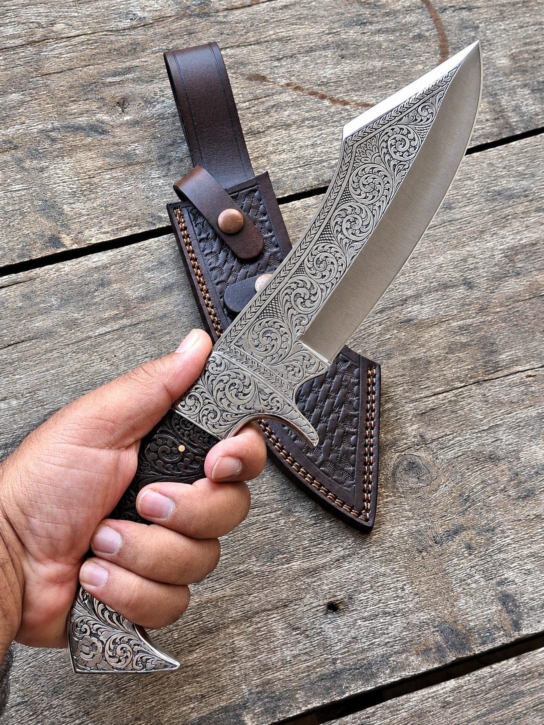 A hand holds a large ornate fixed-blade knife with a silver engraved blade and dark handle, positioned in front of a brown leather sheath on a rustic wooden surface.