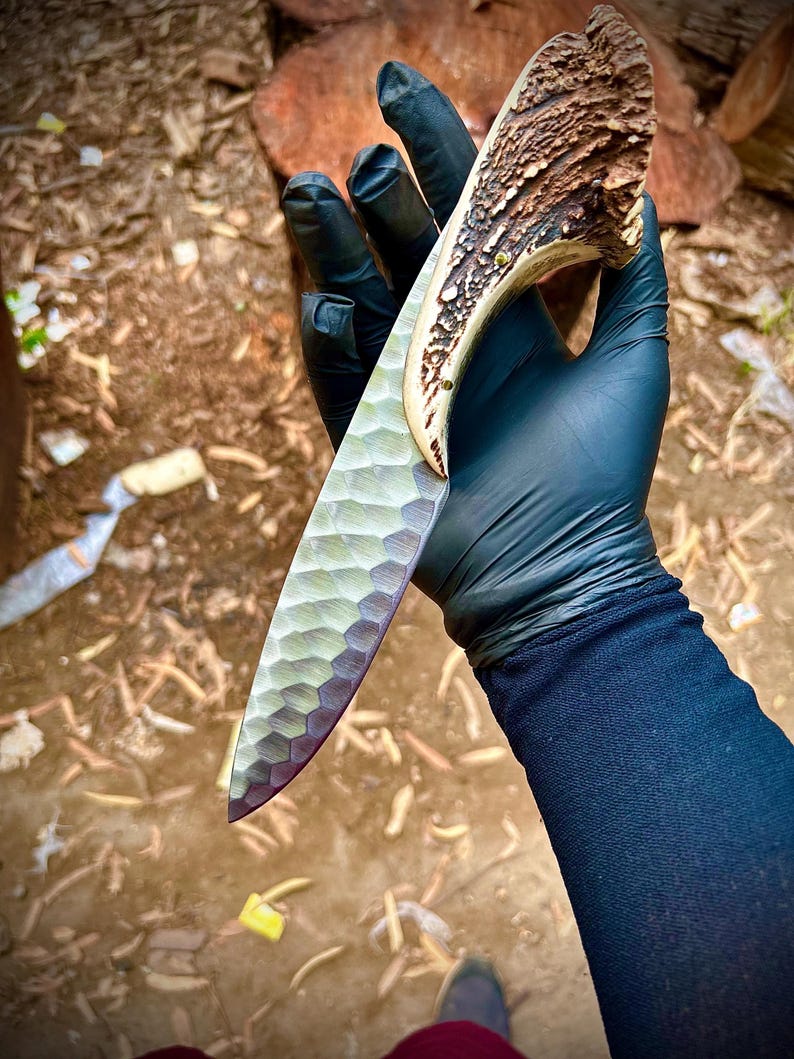 Hand wearing a black glove holding a large knife with a textured blade and wooden handle, on a natural background.