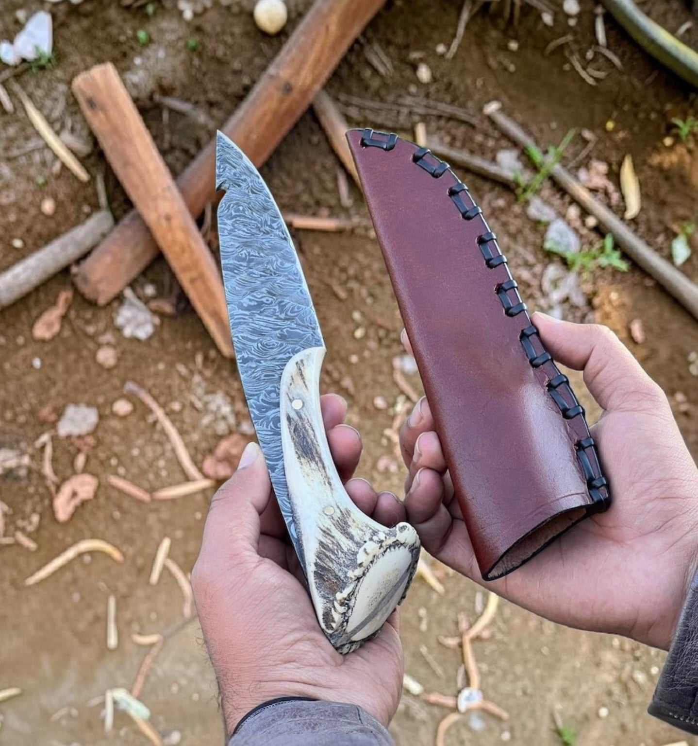 Hand holding a knife with a detailed blade and leather sheath against a natural background.