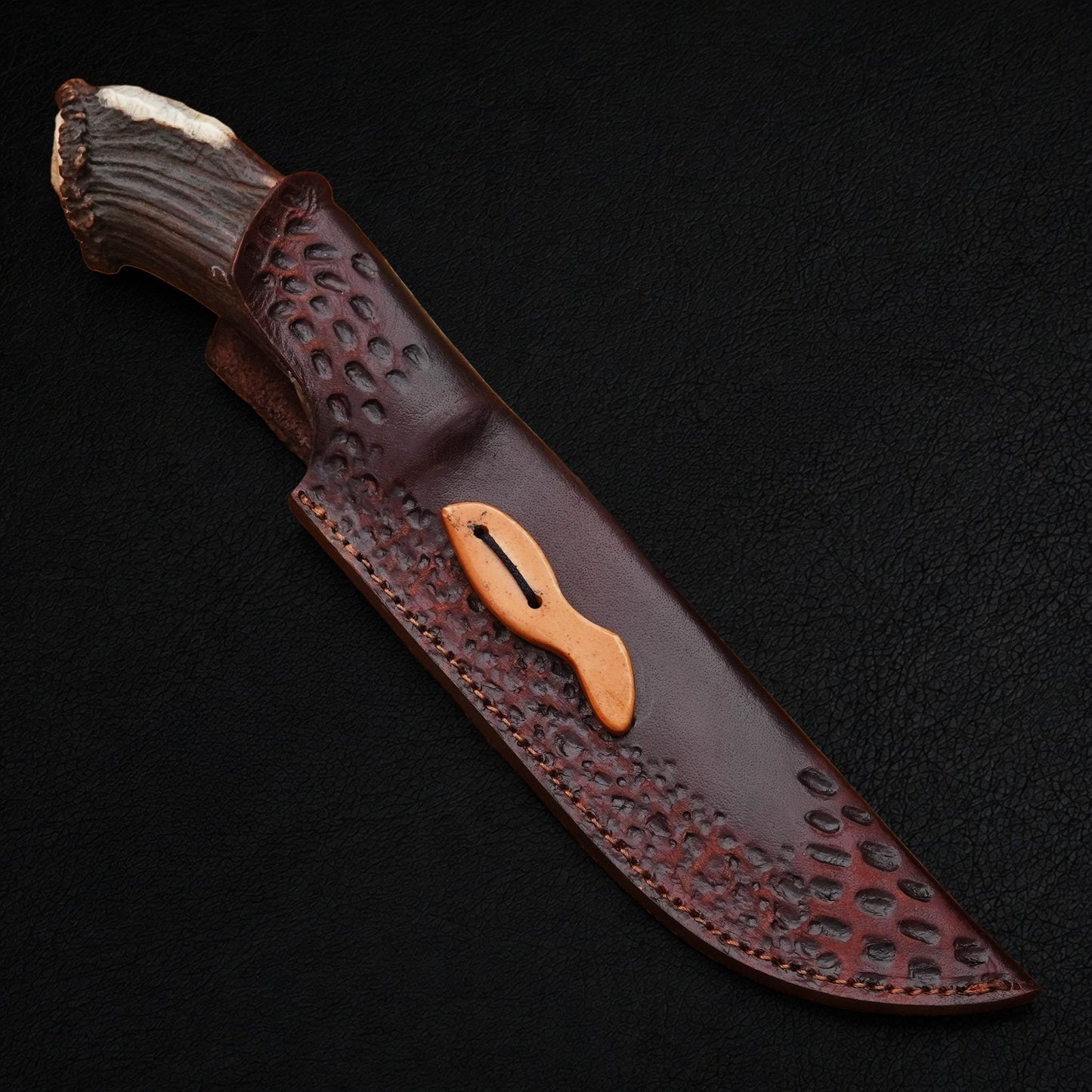 A fixed-blade knife with a textured dark brown leather sheath, featuring a natural antler handle and a light tan wooden guard, displayed diagonally on a black background.