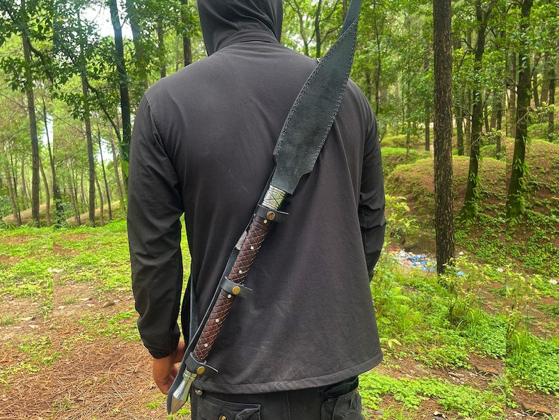 A person viewed from behind wearing a black hooded sweatshirt and standing in a wooded outdoor setting, with a large machete sheathed on their back.