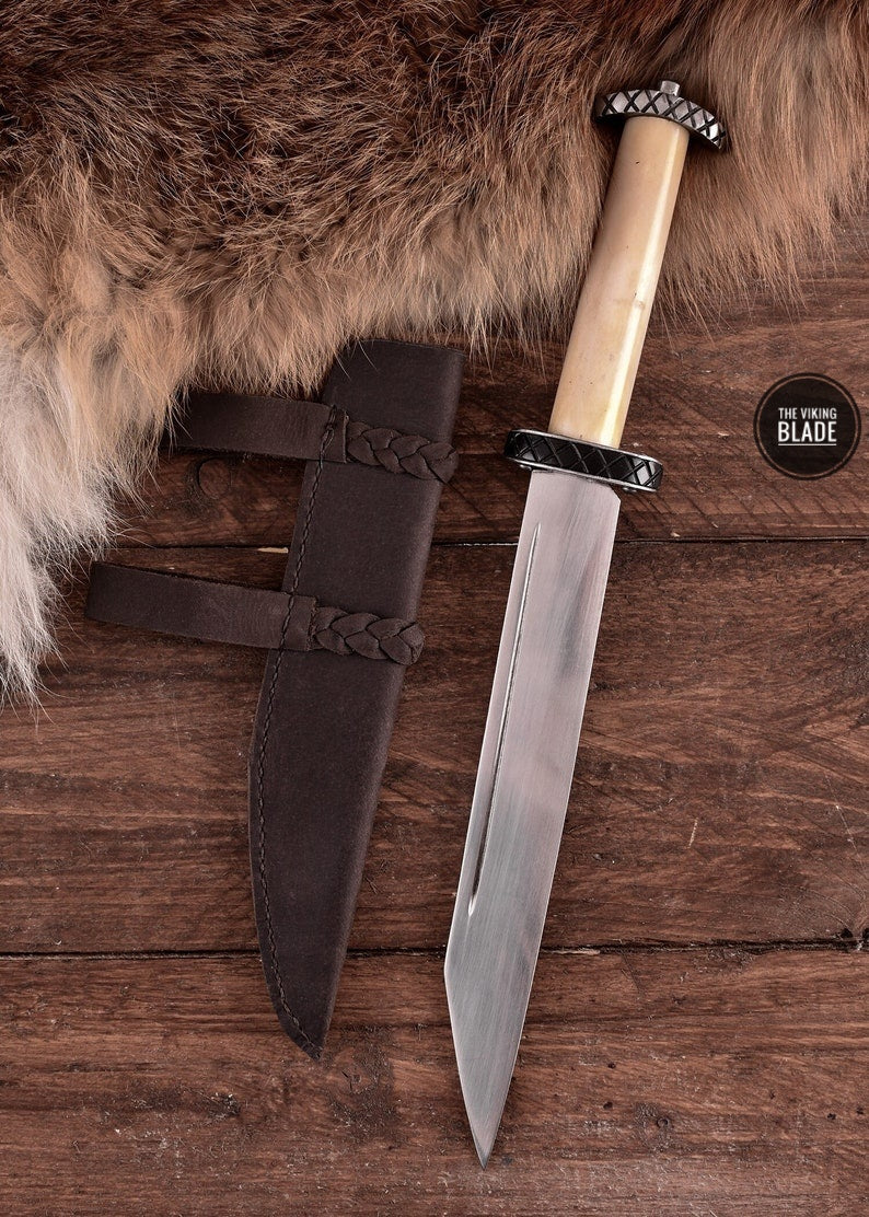 A short sax with a bovine bone handle and a leather sheath, placed on a wooden surface with a furry object in the background.