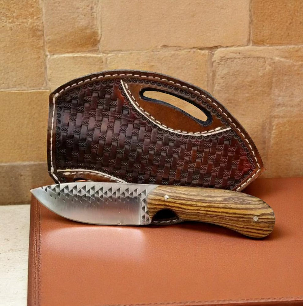 A knife with a high-carbon steel blade and olive wood handle, accompanied by a brown leather sheath with basketweave detailing, displayed against a brick wall backdrop.