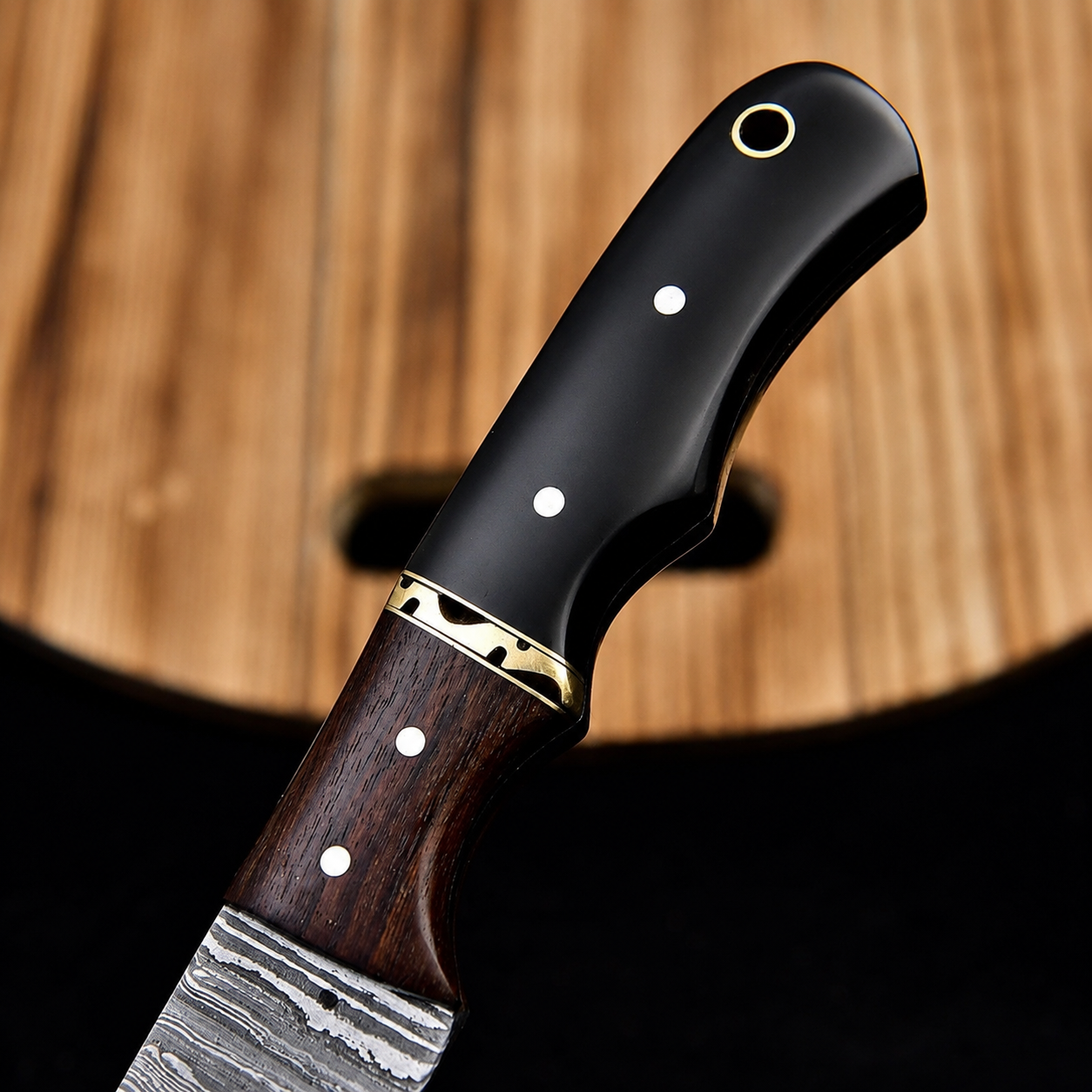 A close-up of a knife handle with a black inlay, white inlays, a gold accent ring, and a Damascus steel blade, set against a blurred wooden background.