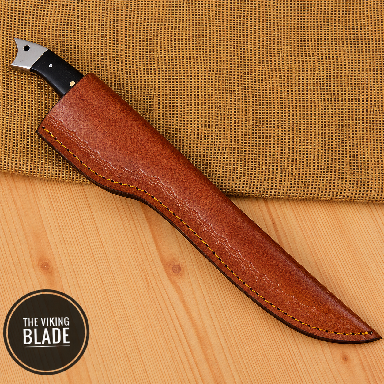 A fixed-blade knife with a black handle and silver bolster, partially sheathed in a brown leather sheath, displayed diagonally on a light wood surface with a burlap cloth background. A circular logo in the bottom-left corner reads 'THE VIKING BLADE'.