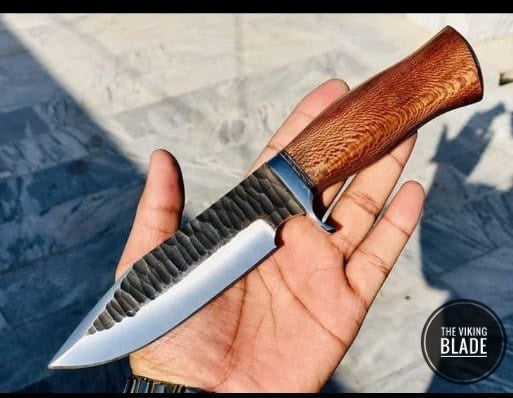Hand forged full tang hunting knife with a wooden handle and a patterned blade, held in a hand, accompanied by a leather sheath.