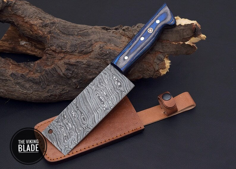 A custom handmade Damascus steel cleaver with a blue wood handle and a patterned steel blade, accompanied by a genuine leather sheath.