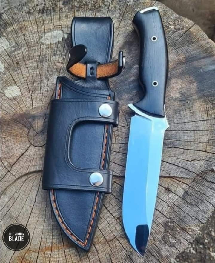 A hand forged J2 steel hunting knife with a black wood handle and a leather sheath, displayed on a wooden surface.