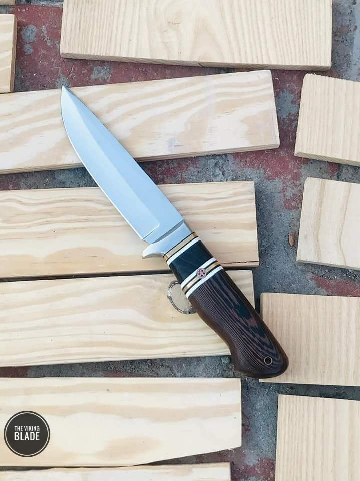 A hand forged J2 hunting knife with a wooden handle and a leather sheath, displayed on a bed of wooden planks.