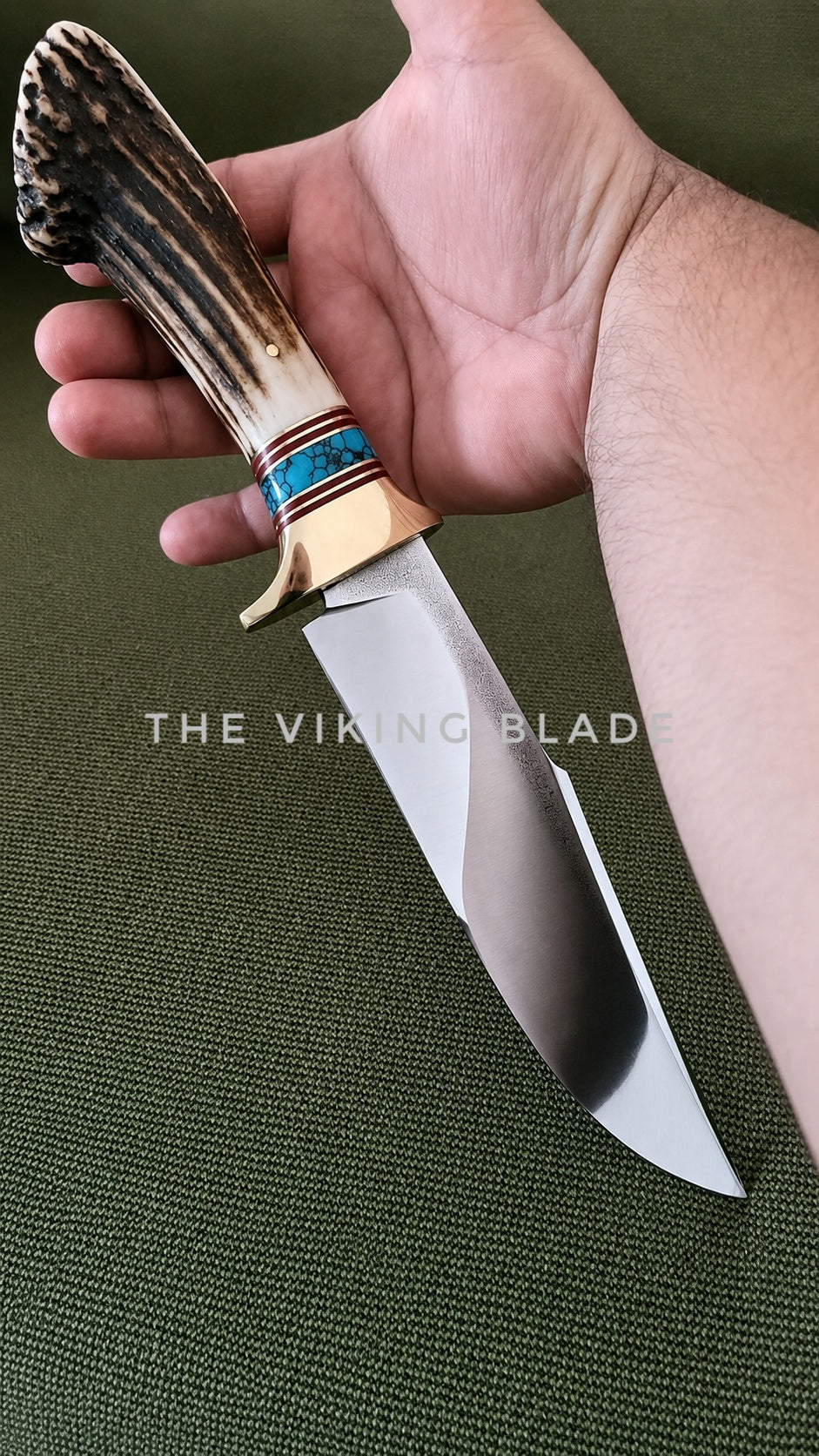 A hand holding a fixed-blade knife with a polished steel blade, a natural antler-style handle, and a turquoise and red inlay near the bolster, set against a dark green textured background. A watermark reading 'THE VIKING BLADE' is visible across the center.