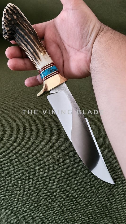 A hand holding a fixed-blade knife with a polished steel blade, a natural antler-style handle, and a turquoise and red inlay near the bolster, set against a dark green textured background. A watermark reading 'THE VIKING BLADE' is visible across the center.