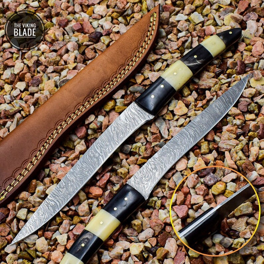 A handmade Damascus steel fillet knife with a buffalo horn and camel bone handle, displayed on a bed of pebbles. The knife has a patterned blade and a smooth, dark handle with a decorative stitching near the guard.