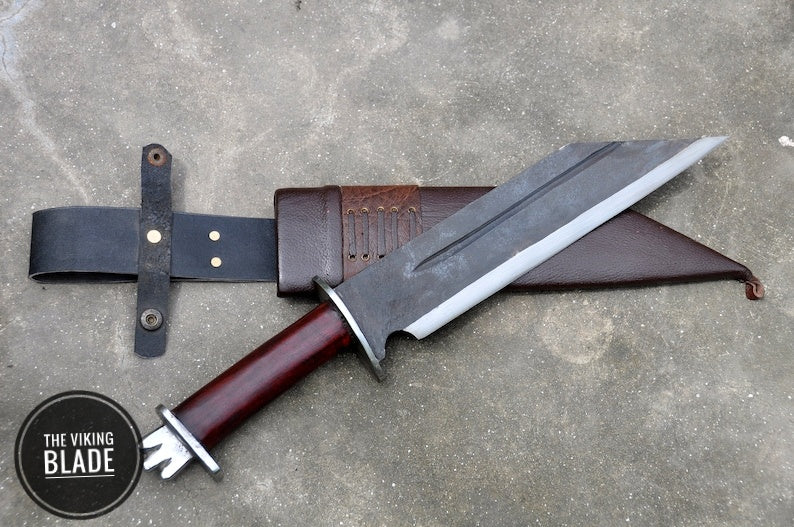 A hand forged seax knife with a polished blade and a brown leather scabbard, laying on a gray surface with a logo 'The Viking Blade' in the corner.