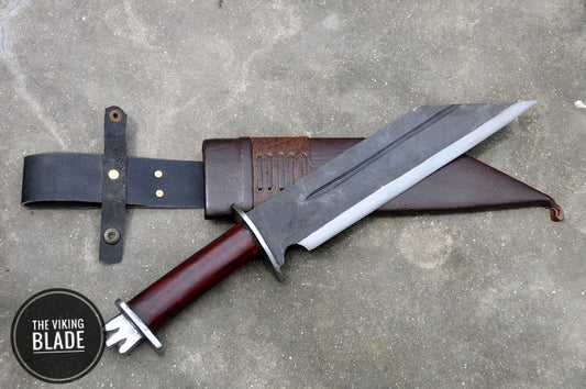 A hand forged seax knife with a polished blade and a brown leather scabbard, laying on a gray surface with a logo 'The Viking Blade' in the corner.