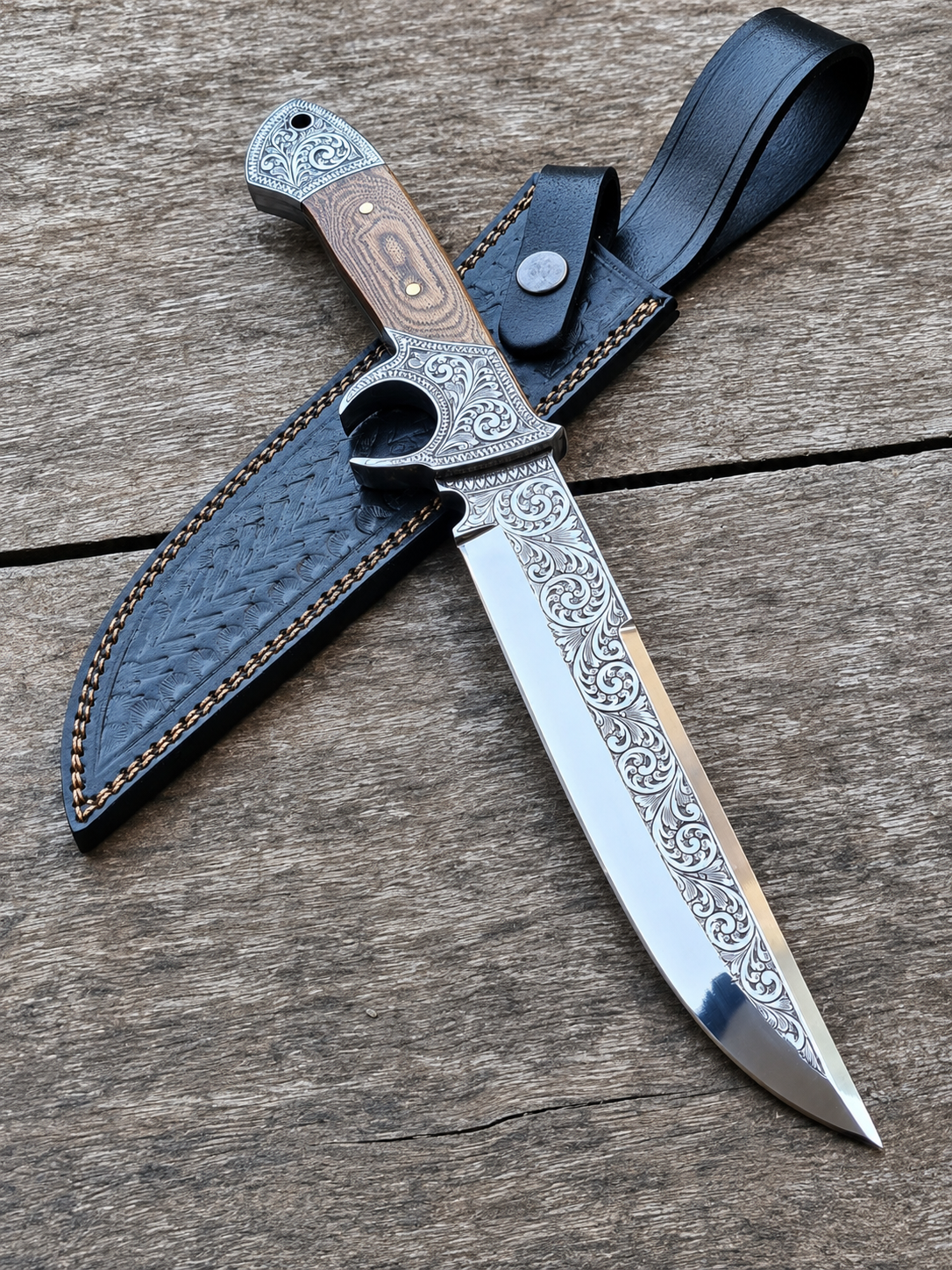 An ornate fixed-blade knife with a patterned steel blade, a wood-and-metal handle, and a black leather sheath, displayed on a rustic wooden surface.