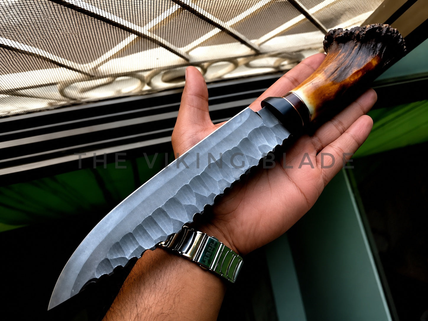 A hand holding a large fixed-blade knife with a hammered steel blade, black bolster, and a polished brown and black horn handle. A watermark reading 'THE VIKING BLADE' is visible across the center.