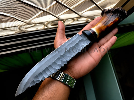 A hand holding a large fixed-blade knife with a hammered steel blade, black bolster, and a polished brown and black horn handle. A watermark reading 'THE VIKING BLADE' is visible across the center.