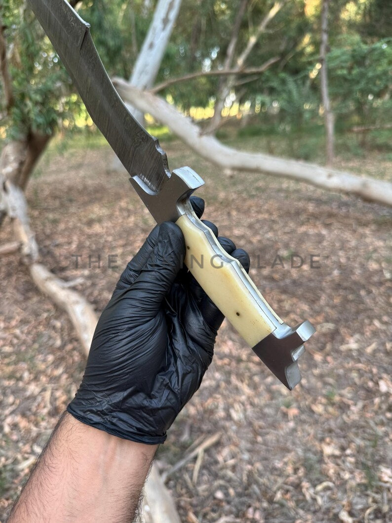 Knife with a black blade and yellow handle held by a gloved hand outdoors.