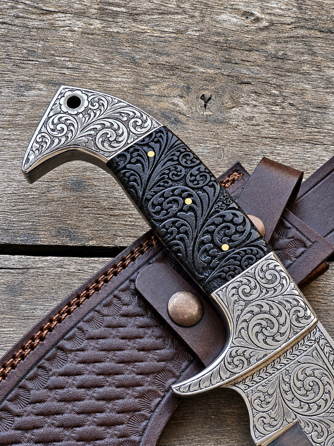 A close-up of a decorative knife with an engraved silver-toned guard and pommel, a black handle with embossed scrollwork and small brass dots, resting on a brown leather sheath over a rustic wooden surface.