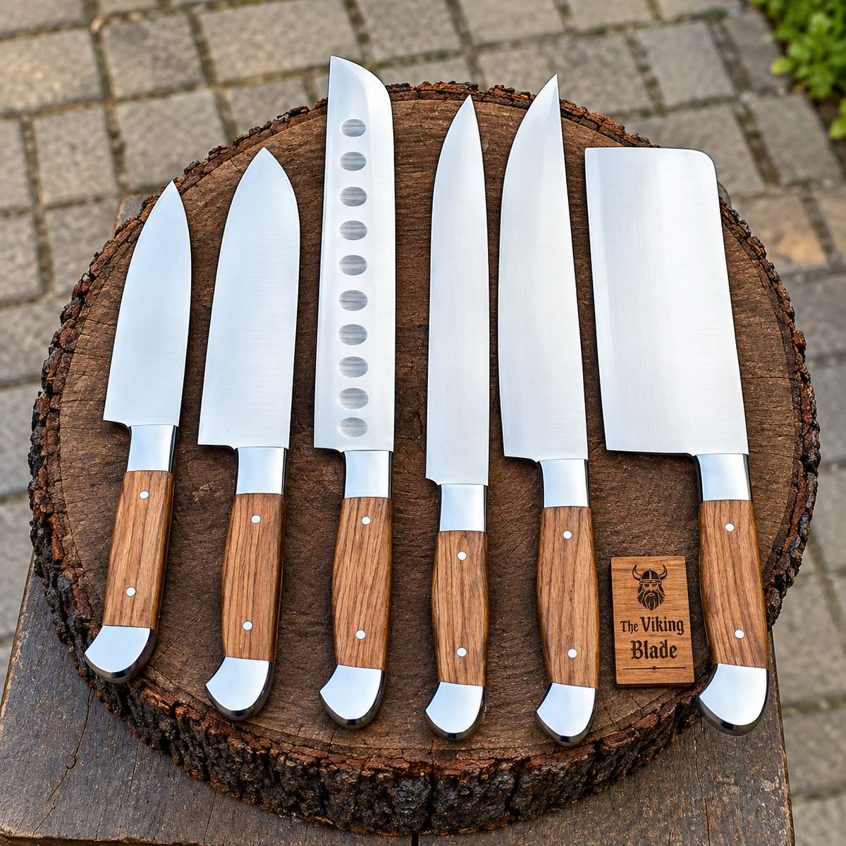 Six stainless steel kitchen knives with wooden handles are arranged on a round wood slice outdoors. The set includes a chef's knife, a santoku-style knife, a utility knife, a slicing knife, a carving knife, and a large cleaver.