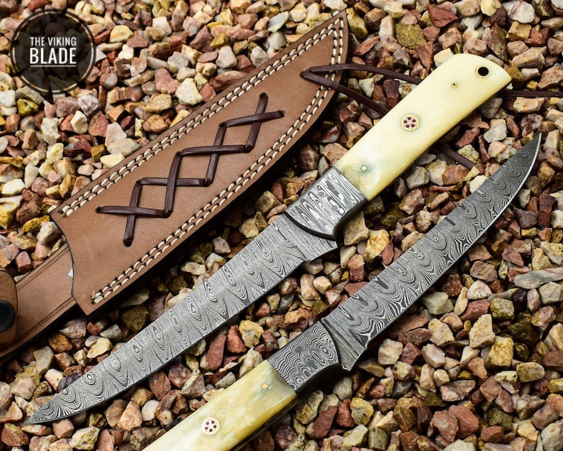 A handmade Damascus steel fillet knife with a camel bone handle, displayed with its blade up and the handle lying flat, set against a bed of pebbles.