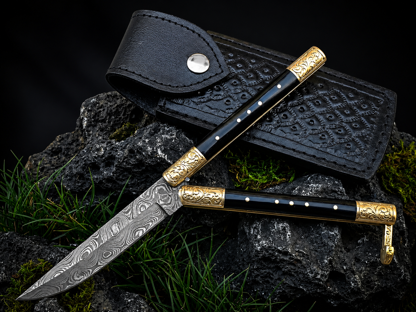 A folding pocket knife with a patterned Damascus steel blade, black handle scales with gold-toned metal accents, displayed next to its black textured leather sheath on dark rocks with green moss.