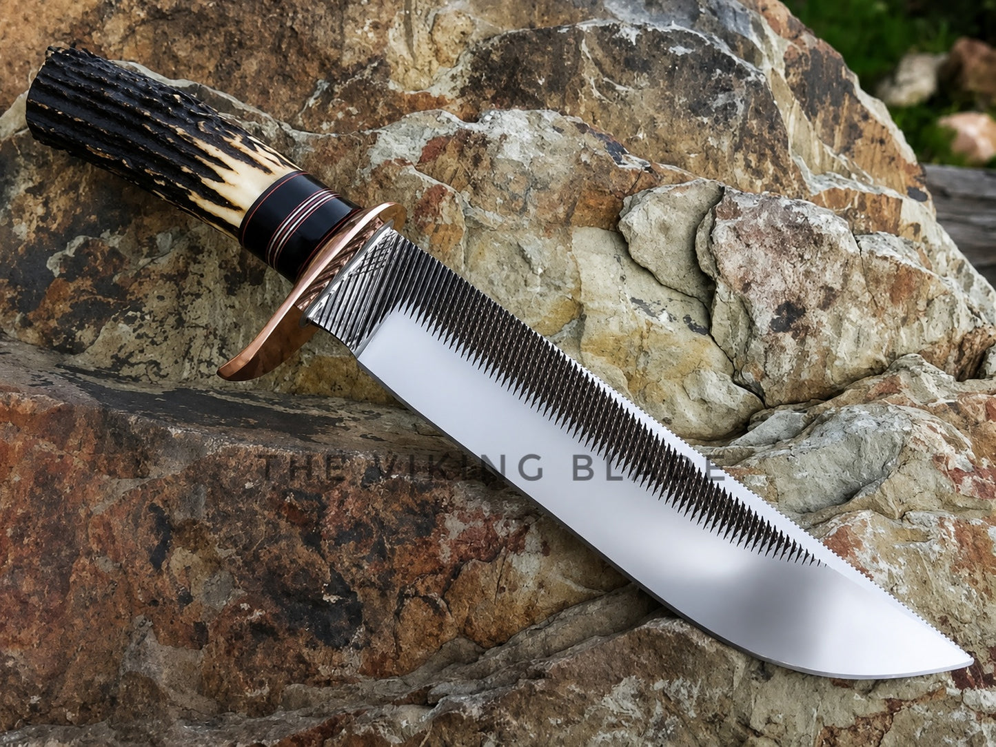 A fixed-blade knife with a textured black and tan handle, a copper-colored bolster, and a serrated silver blade, resting diagonally on a natural rock surface. A watermark reading 'THE VIKING BLADE' is visible across the center.