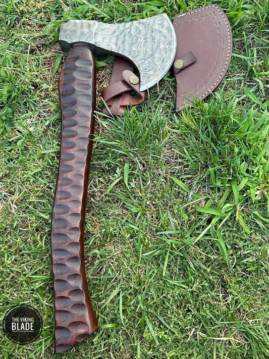 A hand-forged Damascus steel camping hatchet with a brown leather sheath, laying on a grassy surface.