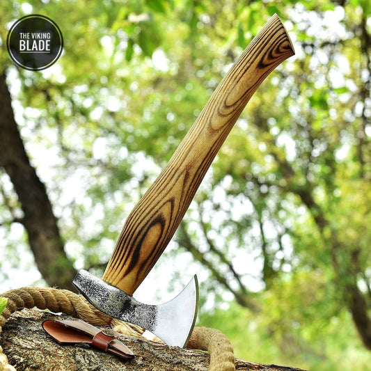 A hand forged hatchet with a wooden handle and a leather sheath, placed on a tree stump with a blurred green background.