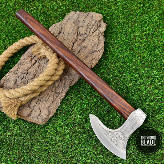 A carbon steel axe with a wooden handle and a leather sheath, placed on a grass-like surface.