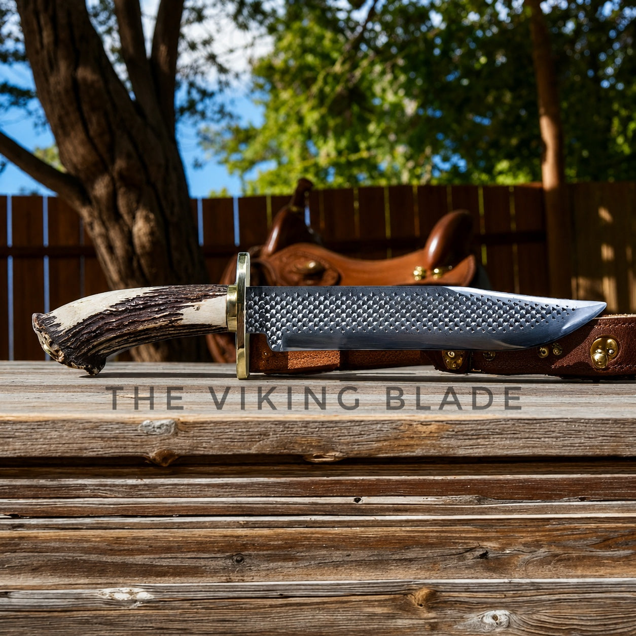 A fixed-blade knife with a textured silver blade, brass guard, and antler-style handle rests on a wooden surface outdoors. A brown leather sheath is partially visible behind it, with a wooden fence and trees in the background.