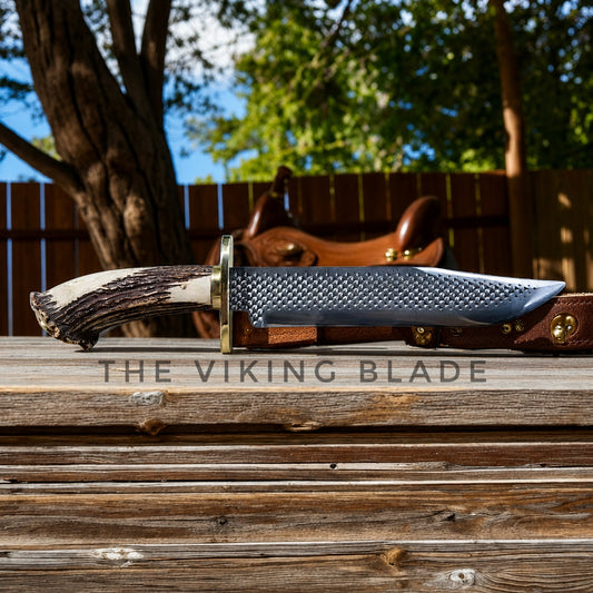 A fixed-blade knife with a textured silver blade, brass guard, and antler-style handle rests on a wooden surface outdoors. A brown leather sheath is partially visible behind it, with a wooden fence and trees in the background.