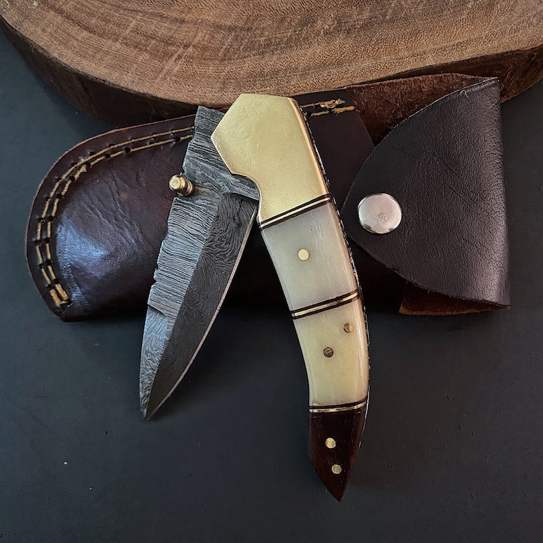 A Damascus steel folding pocket knife with a brown and beige handle, displayed alongside its leather pouch with snap button closure.