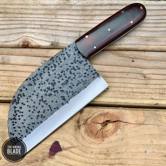 A custom hand forged chopper knife with a patterned blade and brown wooden handle, accompanied by a leather sheath, lying on a wooden surface.