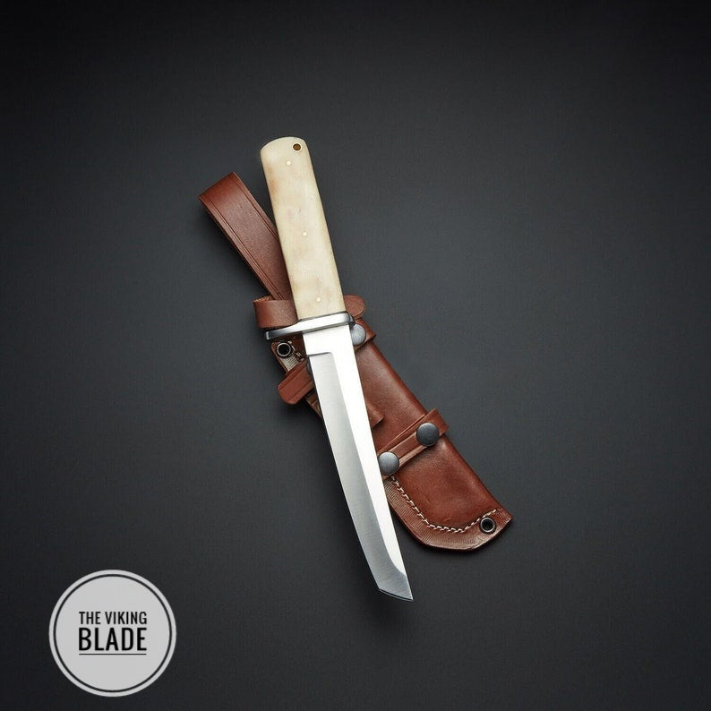 A custom hand forged D2 steel tanto knife with a camel bone handle and a leather sheath, placed on a dark background. The Viking Blade is visible on the knife's handle.