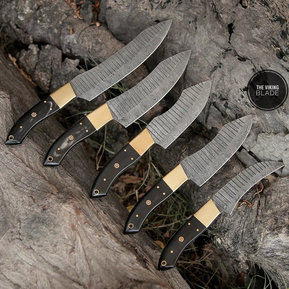 A set of custom hand forged Damascus chef knives with brass bolsters and horn handles, displayed on a wooden surface with a rustic background.
