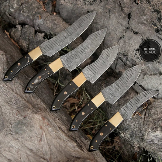 A set of custom hand forged Damascus chef knives with brass bolsters and horn handles, displayed on a wooden surface with a rustic background.