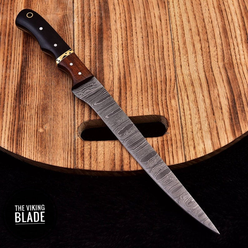 A custom handmade Damascus steel fillet fish chef knife with a brown leather sheath, displayed on a wooden surface.