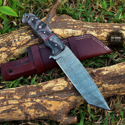 A custom hand forged Damascus steel hunting knife with a Tanto blade design, featuring a brown resin handle and a genuine leather sheath, placed on a natural outdoor surface with wooden logs in the background.