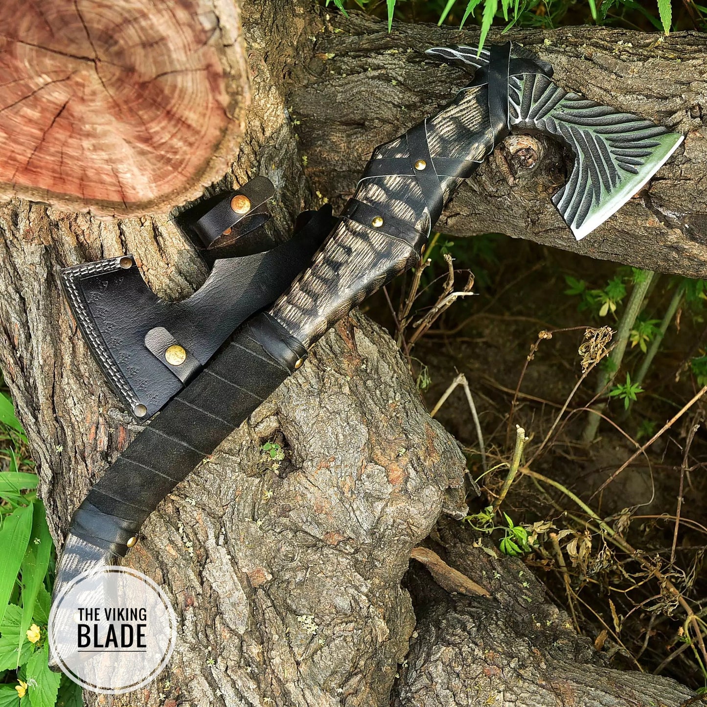 A hand forged Viking axe with a leather sheath, placed vertically against a tree with a natural outdoor background.