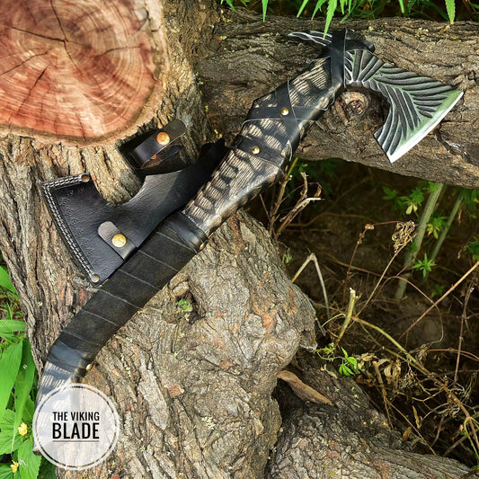 A hand forged Viking axe with a leather sheath, placed vertically against a tree with a natural outdoor background.