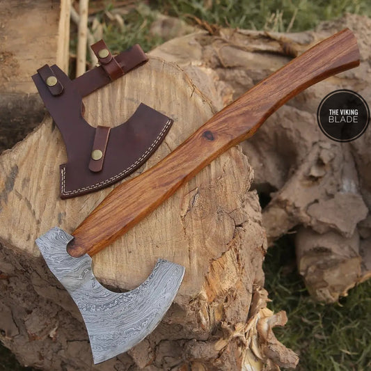 A hand forged Damascus steel axe with a brown leather sheath, placed on a wooden stump with a natural outdoor background.
