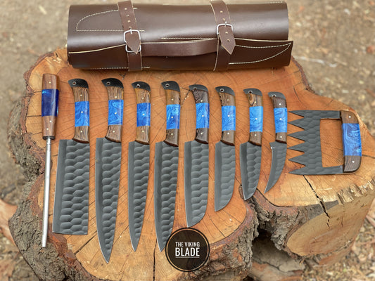 A collection of custom handmade chef knives with black coated blades, displayed alongside a brown leather rolling kit with straps on top. The knives are arranged on a wooden cross-section, showcasing their various handles and blade designs.