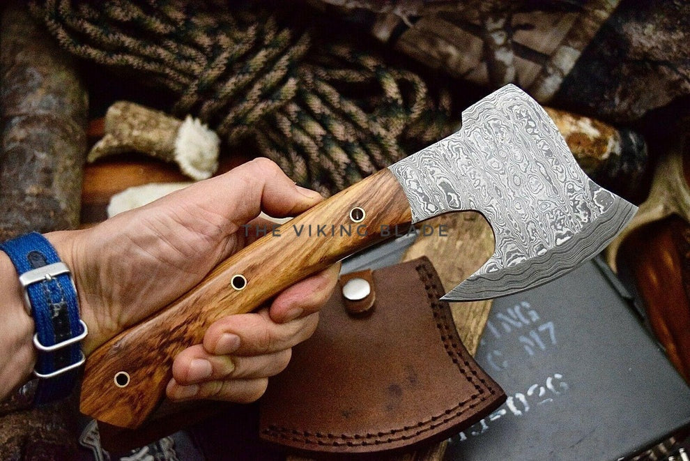 Hand holding a Viking-style axe with a leather sheath, surrounded by natural elements.